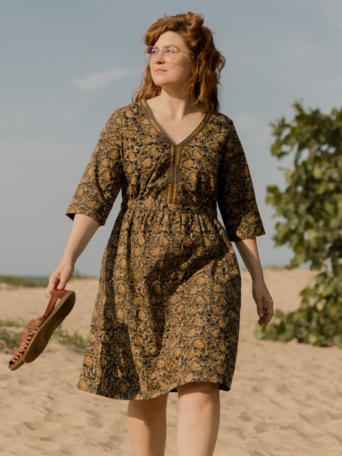 Woman in an olive green  dress walking on a sandy path with trees in the background