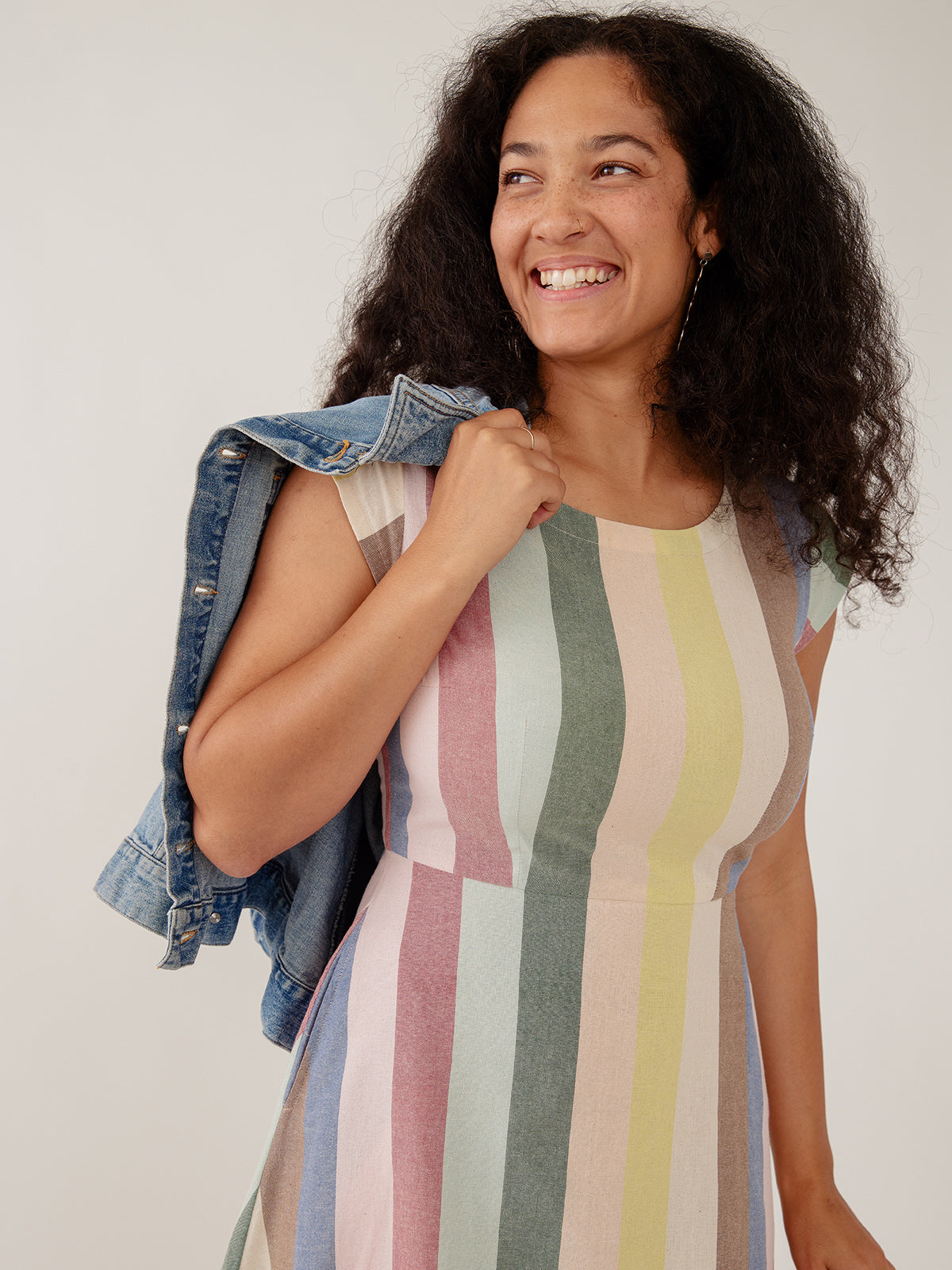 Woman wearing a colorful striped  cotton dress with a denim jacket over her shoulder against a plain background by Mata Traders Sustainable Slow Fashion Brand 