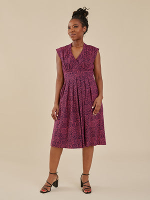 A woman modeling a sleeveless V-neck midi dress with a purple and magenta paisley pattern, paired with black strappy heels, against a solid beige studio background. Mata Traders Fair Trade Dresses