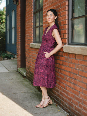 A woman stands against a red brick building wall wearing a sleeveless, knee-length magenta dress with pockets with a tonal floral pattern and nude high-heeled sandals. Mata Traders Natural Fiber Clothing for Women